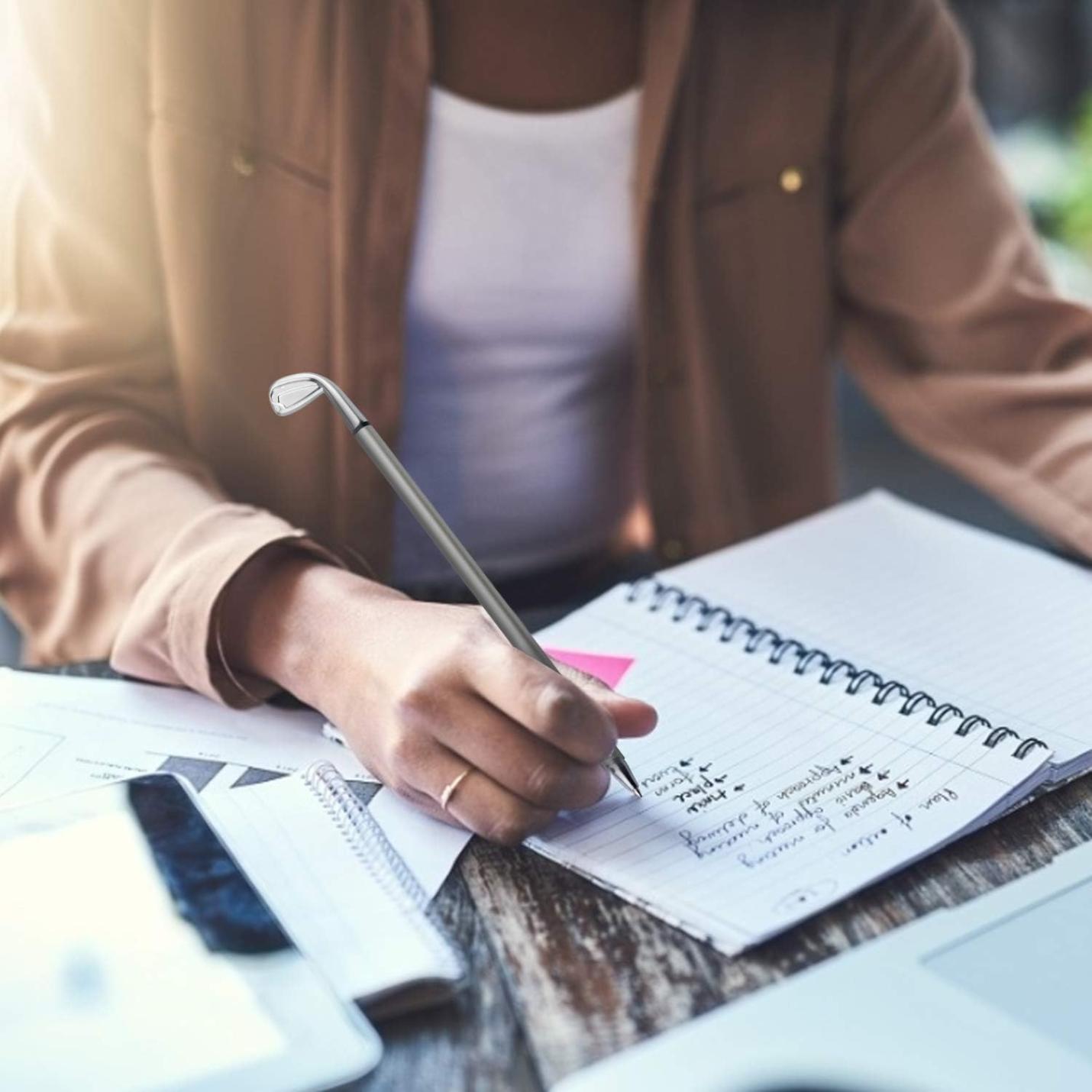 Zin met productnaam: Een persoon die in een notitieboekje schrijft met een golfpennenset van 3 stuks, met diverse papieren en elektronische apparaten op tafel.