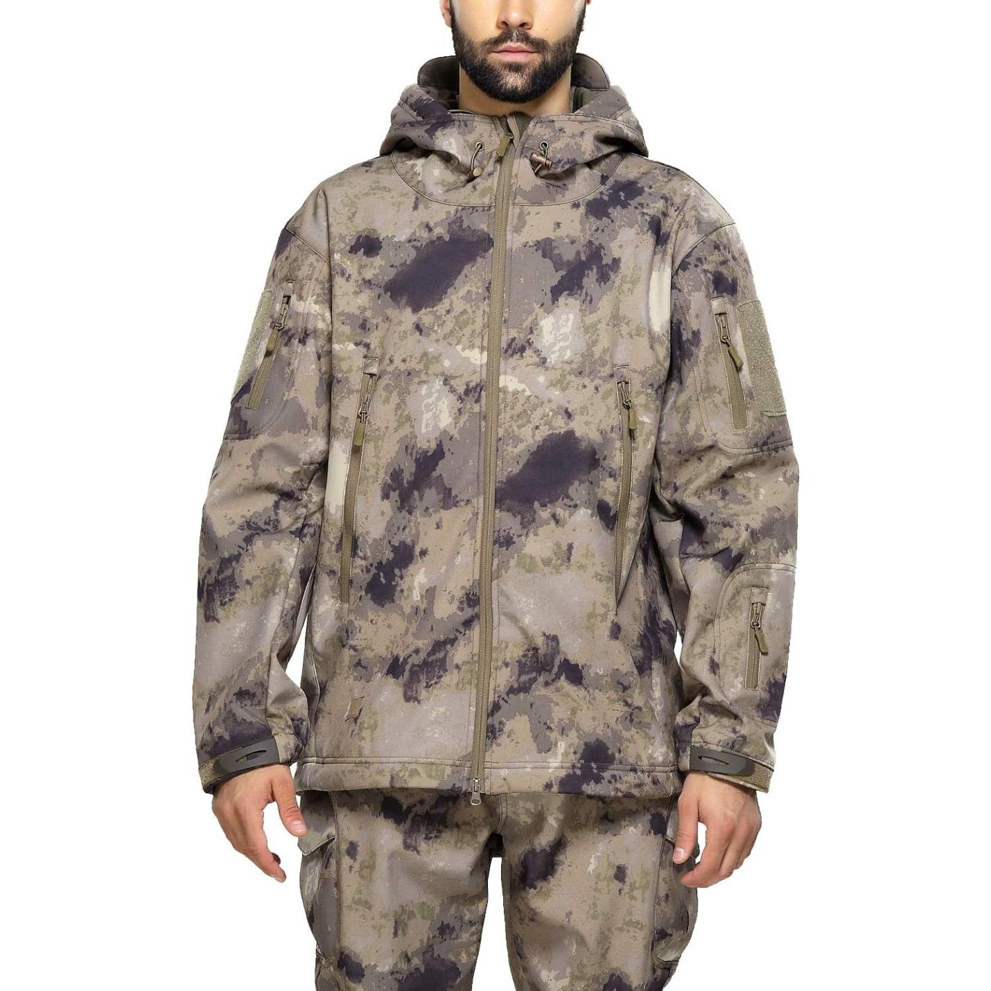 Een man, gekleed in een camouflage tactische heren softshell jas en bijpassende broek, staand met een neutrale uitdrukking.