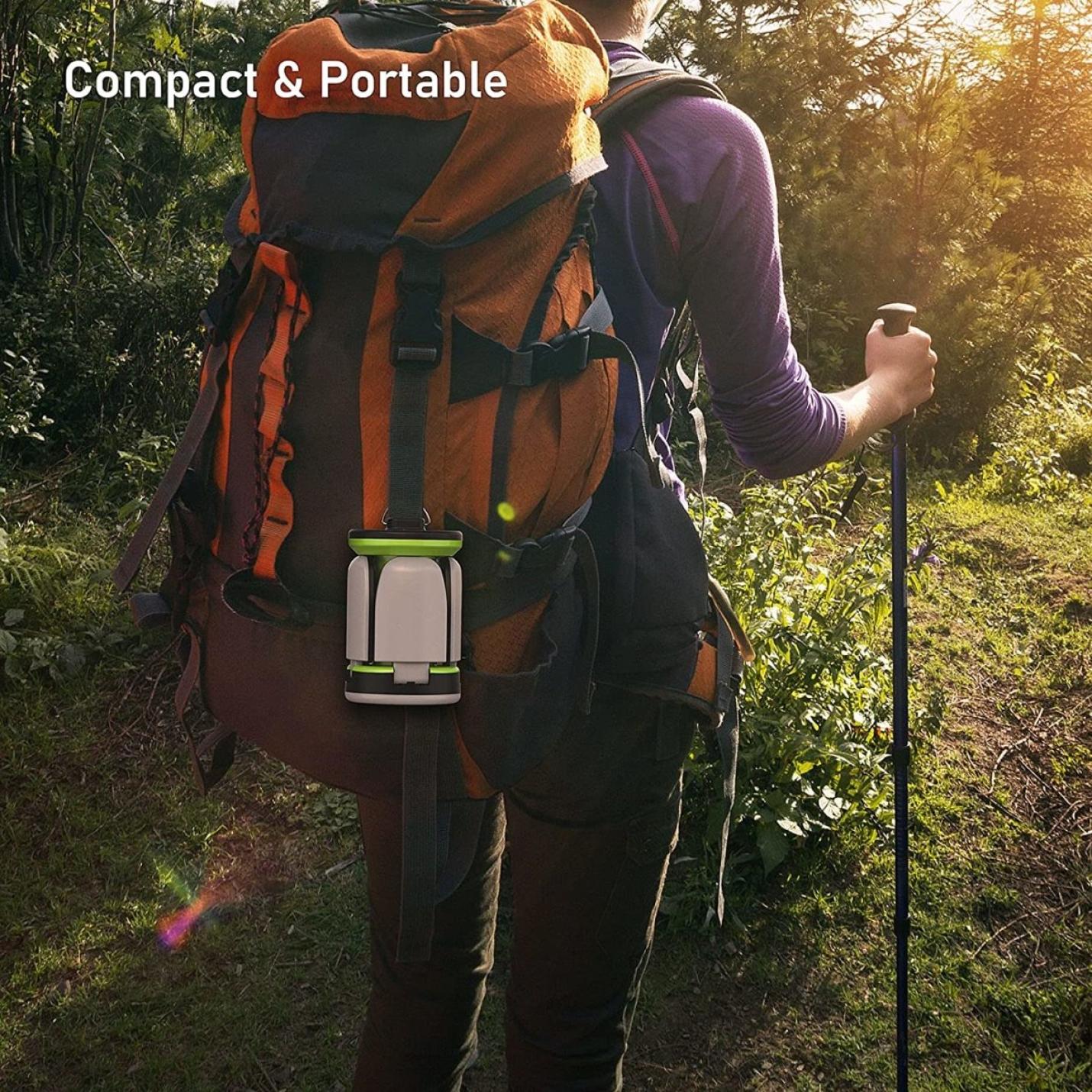 Wandelaar met een oranje rugzak en trekkingstok die door het bos trekt, met een waterdichte en schokbestendige campinglamp voor buitenactiviteiten, samen met andere compacte en draagbare spullen.