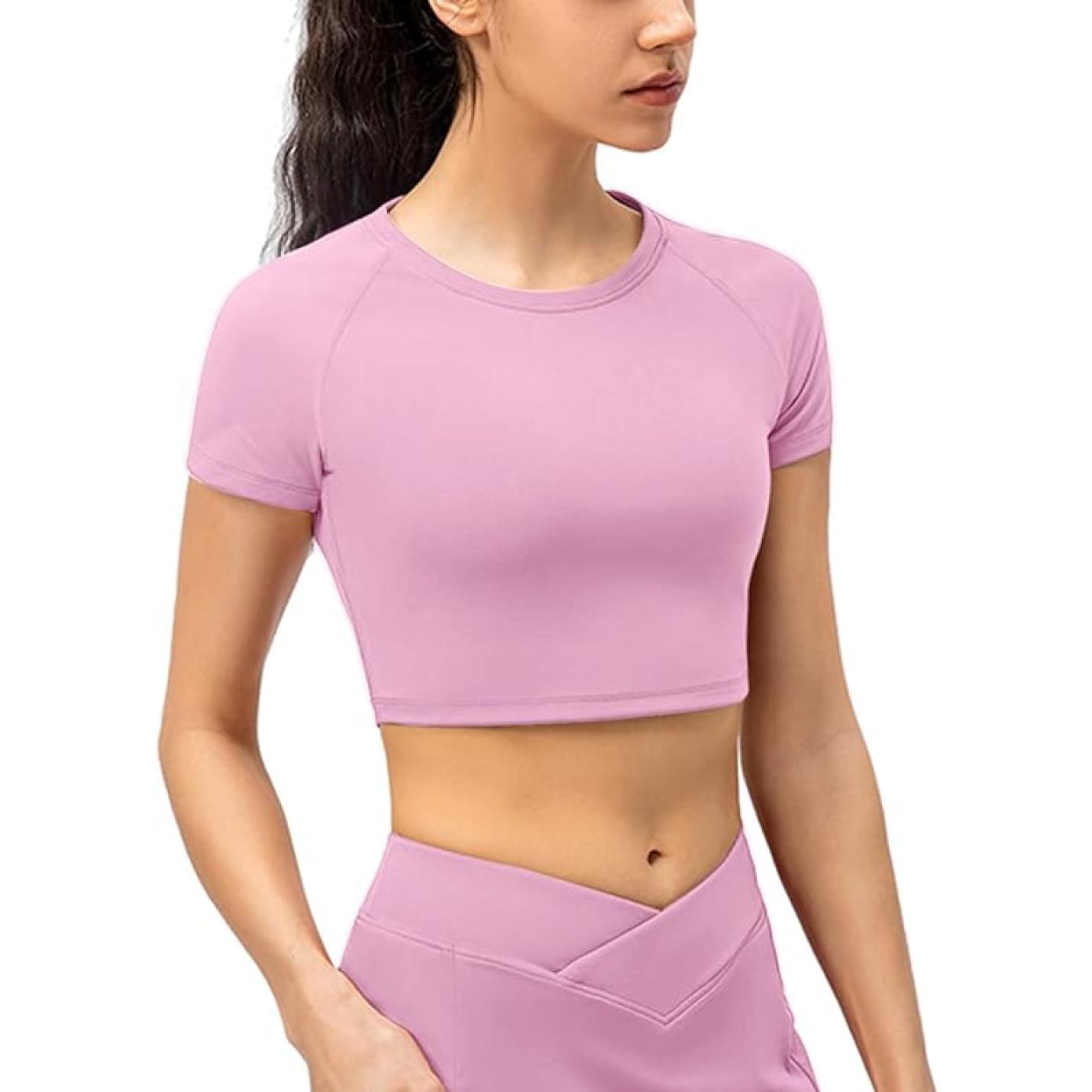 Een vrouw, gekleed in een lichtroze, tijdelijke fitness crop top voor dames en bijpassende legging met hoge taille, staande in een profielhouding.