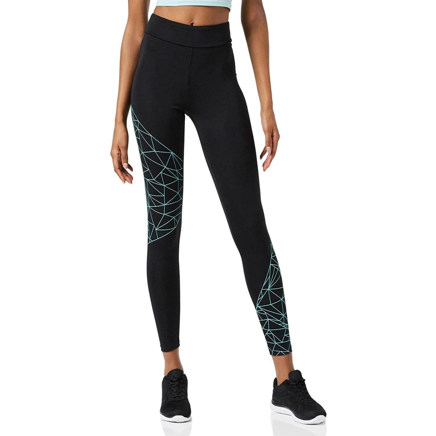 Een vrouw in onze zwarte en teal sportlegging voor dames met geometrisch patroon en zwarte sneakers staat geposeerd tegen een neutrale achtergrond.