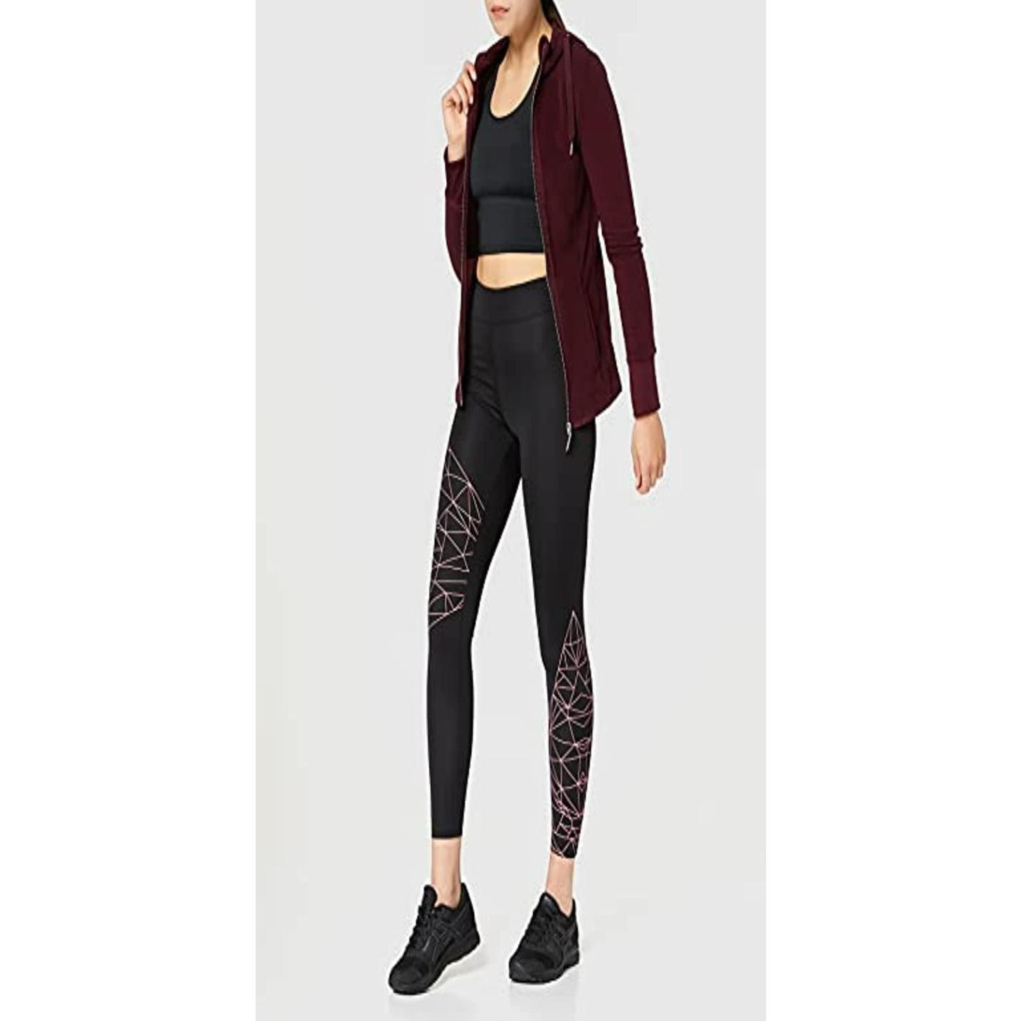 Vrouw in een kastanjebruin jasje, zwarte crop-top en Ultra Stretch STOF zwarte sportlegging voor dames met roze geometrische patronen, zelfverzekerd staande met haar hand op haar heup.